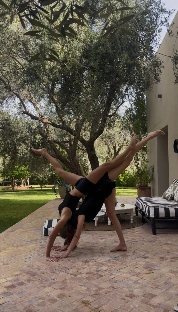 screenshot Two people in black outfits performing a synchronized handstandcartwheel on a brick patio beside a sofa under olive trees in a garden
